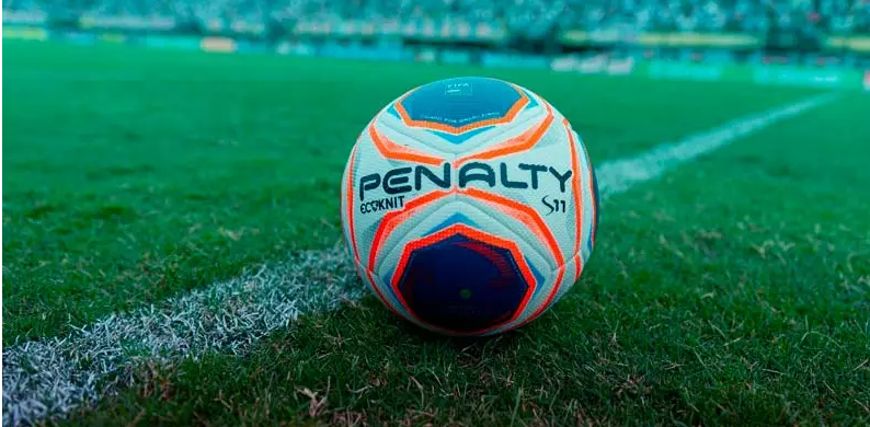 Imagem de uma bola de futebol da marca Penalty no gramado verde de um campo de jogos